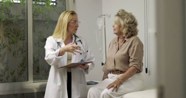 Klinik muayene odasında yaşlı bir kadın hastayla teşhisi tartışırken gülümseyen kadın doktor, her ikisi de sırtüstü serumla sıcak temas kuruyor.