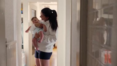 Genç anne nazikçe bebeğini kucaklıyor, koridorda dikilirken bebeğin başını öpüyor. Yansıma yakınlardaki bir aynaya yansıdı.