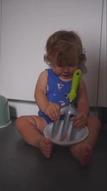 Küçük çocuk mutfak zemininde çatal bıçak tutarak ve kâseyle oynayarak çocukluk yıllarında merak, keşif ve eğlenceli keşifler yapıyor.