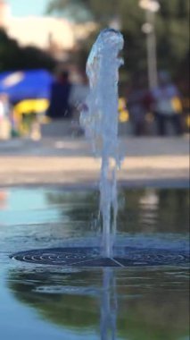 Şehir çeşmesinden yükselen su püskürtüsü yüzeyde dalgalanan yansıma ve şehir meydanının bulanık arkaplanı tazelik ve hareket çağrıştırıyor