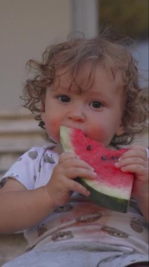 Kıvırcık saçlı çocuk elinde büyük bir karpuz dilimi, yüzü ve gömleği meyve suyuyla dağılmış, açık havada tatlı meyvenin tadını çıkarıyor.