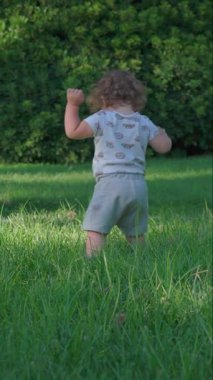 Kıvırcık saçlı çocuk parkta uzun yeşil çimenlerde yürüyor, açık havayı düzenli adımlarla ve erken çocukluk merakıyla keşfediyor.