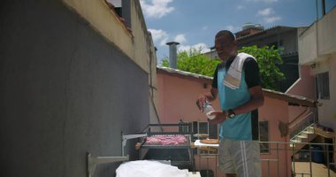 Bir siyah adam açık havada et tatlandırıyor, Latin Amerika 'nın arka bahçesinde konut binalarıyla barbekü hazırlıyor.