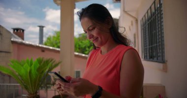 Dışarıda akıllı telefonu tutan, üzgün ve düşünceli görünen, arka planda yeşillik olan sıradan bir mesken ortamının yanında duran bir kadın.