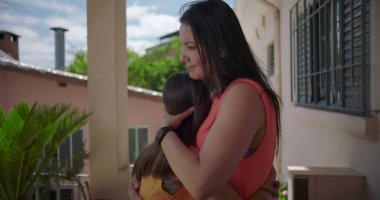 Anne, Latin Amerika 'da bir balkonda kızını kucaklıyor. Arka planda yeşillik ve pastel konut binalarıyla sevgi dolu bir anın tadını çıkarıyor.