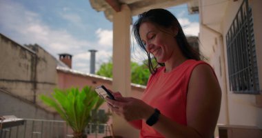 Dışarıda elini kaldıran ve akıllı telefon mesajına heyecanla tepki veren, neşeli bir sürpriz gösteren, arka planda bitkilerle sıradan bir mesken ortamında duran bir kadın.