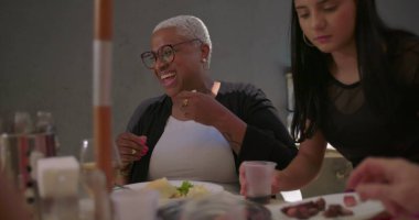 Arka bahçede öğle yemeği yerken neşeyle gülen bir kadın, elinde peçeteyle yemek yiyor ve masada aile ve arkadaşlarla kaynaşıyor.