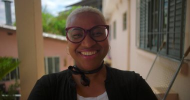 Sıcak bir tebessümle Afrika kökenli Latin bir kadın güneşli bir terasta, kollarını kavuşturmuş, huzur dolu bir Latin Amerika yerleşim yerine güven ve gurur yayıyor.