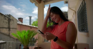 Dışarıda akıllı telefonu tutan bir kadın, hayal kırıklığı ya da inançsızlık içinde alnına vuruyor, arka planda yeşillik olan sıradan bir yerleşim yerinde duruyor.