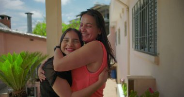 Anne kızı kucaklıyor, hem içten gülümsüyor, hem de yakınlığını ve mutluluğunu ifade ediyor, arka planda bitkiler olan Latin Amerika yerleşim bölgesinde.