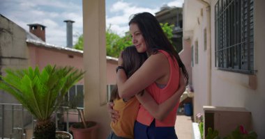 Anne, Latin Amerika 'nın balkonunda sıcak ve duygusal bir anda kızına sarılıyor. Etrafı bitkilerle ve pastel renkli konutlarla çevrili.
