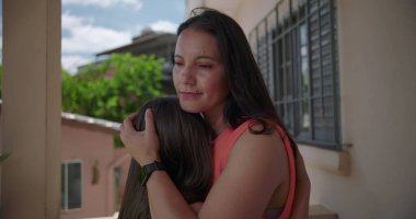 Arka planda yeşil ve pastel binalar olan Latin Amerika 'nın balkonunda kızını kucaklayan bir annenin yakın çekimi.