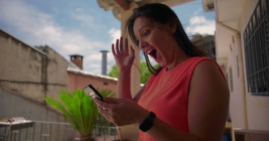 Dışarıda elini kaldıran ve akıllı telefon mesajına heyecanla tepki veren, neşeli bir sürpriz gösteren, arka planda bitkilerle sıradan bir mesken ortamında duran bir kadın.