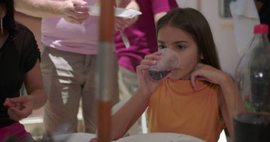 Aile yemeğinde bir bardaktan soda içen genç bir kız, odaklanmış ve rahatlamış, açık hava toplantısı sırasında konuşmalar ve yemekle çevrili.