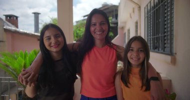 Güney Amerika 'nın sıcak bir semtinde, verandada duran iki kızıyla birlikte gülümseyen anne, birlikte bir aile anının tadını çıkarıyor, yeşilliklerle çevrili.