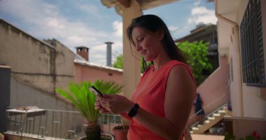 Dışarıda akıllı telefonu tutan, mesaj okurken gülümseyen, meşgul ve odaklanmış görünen, arka planda yeşillik olan sıradan bir mesken ortamında duran bir kadın.