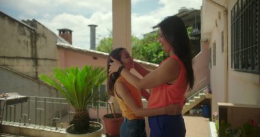 Anne, Güney Amerika 'da bir verandada gülümserken, küçük kızlarının yüzünü nazikçe tutuyor. Sevgisini ve aile bağını gösteriyor..