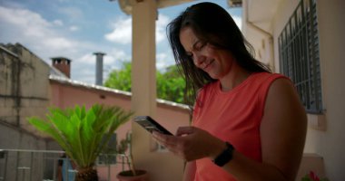 Dışarıda akıllı telefonu tutan bir kadın, hayal kırıklığı ya da inançsızlık içinde alnına vuruyor, arka planda yeşillik olan sıradan bir yerleşim yerinde duruyor.