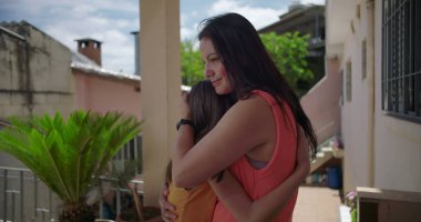 Anne, Latin Amerika 'nın balkonunda sıcak ve duygusal bir anda kızına sarılıyor. Etrafı bitkilerle ve pastel renkli konutlarla çevrili.