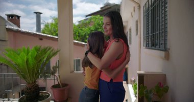 Anne, Latin Amerika 'nın balkonunda sıcak ve duygusal bir anda kızına sarılıyor. Etrafı bitkilerle ve pastel renkli konutlarla çevrili.
