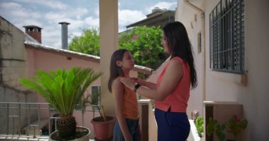 Anne, Latin Amerika 'da bir balkonda kızını kucaklıyor. Arka planda yeşillik ve pastel konut binalarıyla sevgi dolu bir anın tadını çıkarıyor.