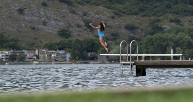 Çocuk 800 fps yavaş çekimde göle dalış platformundan atlıyor, yaz eğlencesinin heyecanını ve maceraperest ruhunu yakalıyor manzaralı bir arka planla.