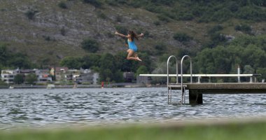 Çocuk 800 fps yavaş çekimde göle dalış platformundan atlıyor, yaz eğlencesinin heyecanını ve maceraperest ruhunu yakalıyor manzaralı bir arka planla.