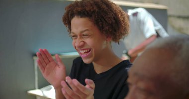 Genç bir çocuk aile toplantısı sırasında neşeyle gülüyor, mutluluk ve kaygısız enerjiyi ifade ediyor, farklı bir aile ortamında canlı bir bağ kuruyor.