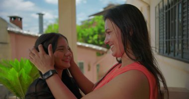 Anne, Güney Amerika 'da yeşillik içinde bir Latin yerleşim yerinde kızlarını nazikçe kucaklıyor, hem sıcak bir şekilde gülümsüyor hem de sevgisini ve bakımını ifade ediyor..