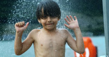 Mutlu çocuk su sıçratarak eğleniyor, ellerini kaldırıyor, açık havada, bulanık doğal bir zemin ile.