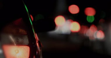 Parlak kırmızı stop lambalı arabanın arkası, uzun bulanık trafik ışıkları şeritleri ile gece caddesinin geniş görüntüsü karanlığa uzanıyor, kentsel gecikmeyi çağrıştırıyor.