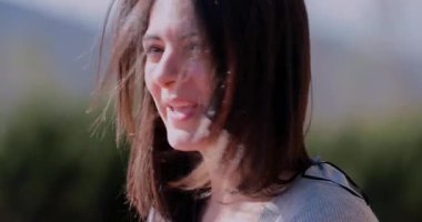 Latin Latin kadın gün ışığında gevşek saçları ile gülümsüyor, açık hava ortamında memnuniyet ifadesiyle yan tarafa bakıyor bulanık doğal arka plan ile