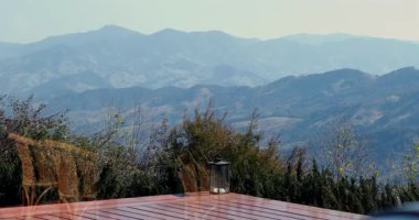 Ahşap güverteden dağ manzarası, uzak tepeler ve ağaçlar, sandalye ve fenerin cam yansıması berrak gün ışığı altında manzarada görünür.