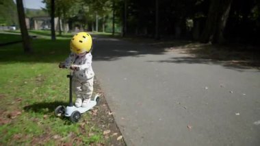 Sarı kask takmış, park yolunda scooter süren çocuk, çimlere yuvarlandıktan sonra yavaşlıyor ve duruyor, kaldırıma dönüp sürmeye devam etmeden önce kısaca sıkışıyor.