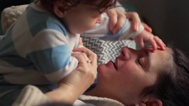 Anne kanepede yatarken bebekle oynuyor. Evdeki kahkahalarla neşe dolu bir etkileşim.