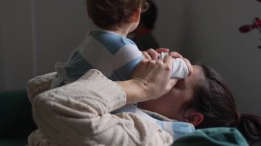 Anne bebekle oynarken sevinçle gülüyor, kanepede bebek ellerini tutuyor, sıcaklığını, şefkatini, mutluluğunu ve duygusal bağını gösteriyor.