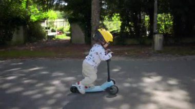 Eğri park yolunda mobiletle gezen çocuk sarı kask takıyor, eğlenceli ve kararlı bir ifade takınıyor, açık havada, neşeli bir çocukluk macerası sürüyor.