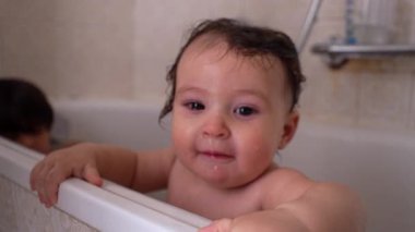 Gülümseyen bebek küvette durmuş, kenarlarını tutarak neşeli bir ifadeyle ve banyo sırasında ıslak saçlarla.