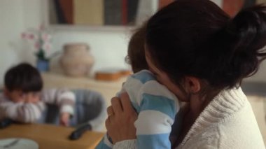 Anne, çocuğu kucağında büyük kardeşiyle yemek masasında otururken bebeğini başından öpüyor.