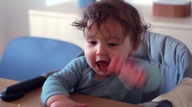 Yüksek sandalyede oturan kıvırcık saçlı çocuk, tuhaf bir ifadeyle masaya bakıyor, elleri önde dinleniyor, evde çocukluk anıları yaşıyor.