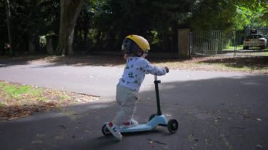 Park yolunda scooter süren bir çocuk sarı başlık takıyor, açık havada keyifli bir an odaklanmış bir ifade ile yol boyunca süzülüyor, yeşillik içinde.