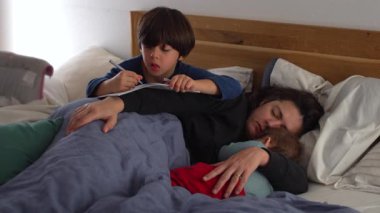 Anne kucağında bebekle yatakta dinlenirken, büyük çocuk onun yanında yazıyor, günlük yaşamında samimi aile bakımı, bağ kurma ve birliktelik anları