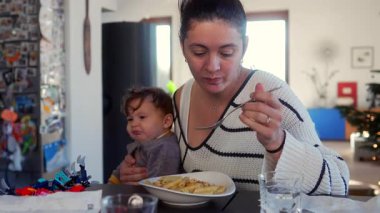 Anne evde yemek saatinde bebeği kucağında tutarken makarna yiyor.
