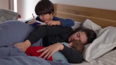 Anne yatakta dinleniyor, bebeğe sarılıyor. Büyük çocuk yanında yazıyor. Sessiz bir şefkat anı, günlük ebeveynlik aşkı ve yakın aile birlikteliği.