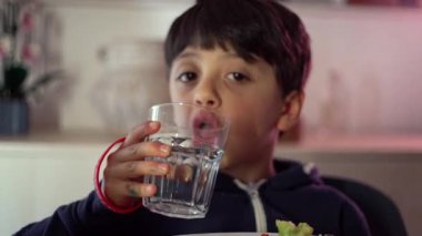 Evde yemek saatinde yemek masasında temiz bardaktan su içen genç bir çocuk.