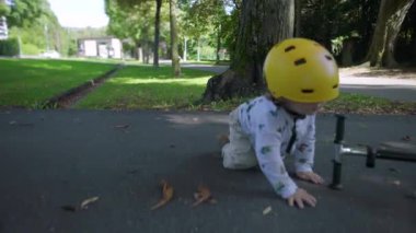 Sarı kasklı bir çocuk kaldırım parkında scooter 'dan aşağı düşüyor, çocuk dışarıda oynarken tökezliyor ve aniden yere düşüyor.
