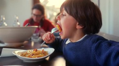 Genç çocuk, arka planda annesiyle yemek yerken mutfak masasında çatal makarna yemek için ağzını kocaman açıyor..