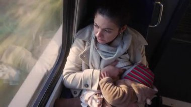 Genç anne, trende bebeğini emzirirken pencereden dışarı bakar, yolculuk sırasında sakin bir yansıma ve beslenme anı, anne sevgisi ve bağ.