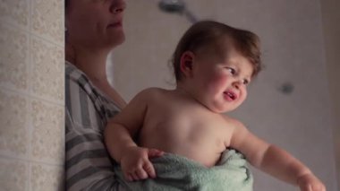 Anne banyodan sonra havluya sarılmış bebeği tutarken, çocuk merakla, samimi aile buluşları, şefkat ve annelik bakımı anlarına bakıyor..