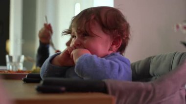 Hüsrana uğramış mavi elbiseli çocuk yüksek sandalyede oturuyor ve evde aile yemeği sırasında dışarı çıkmaya can atıyor.